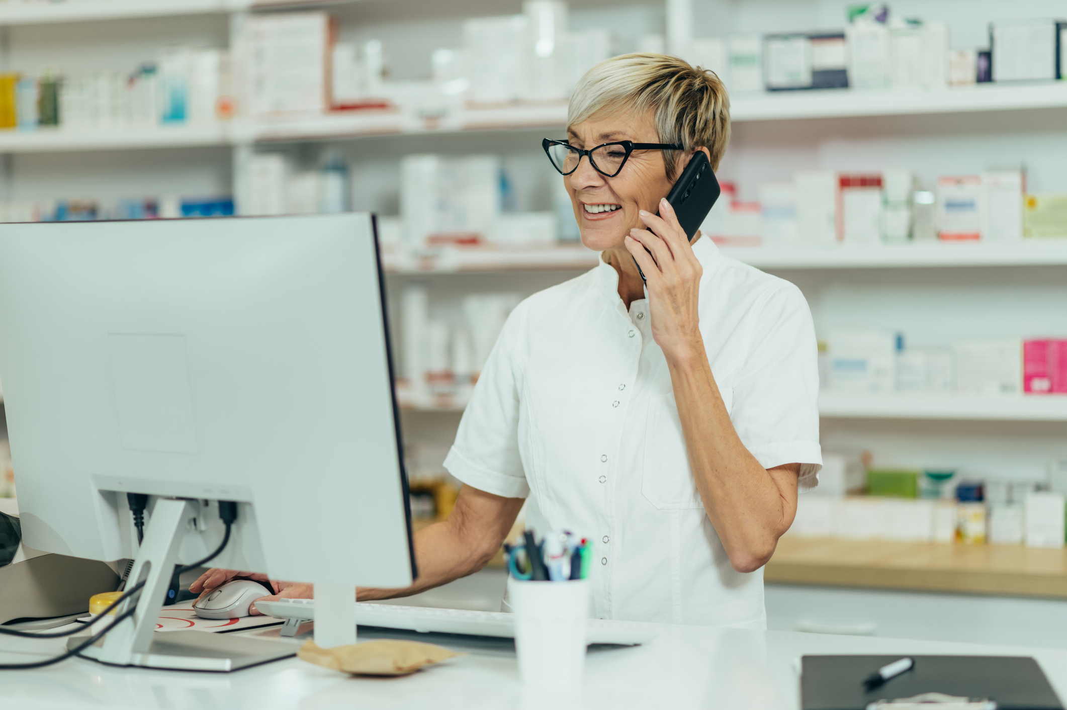 An older, female pharmacist talks on the phone while looking at a computer.