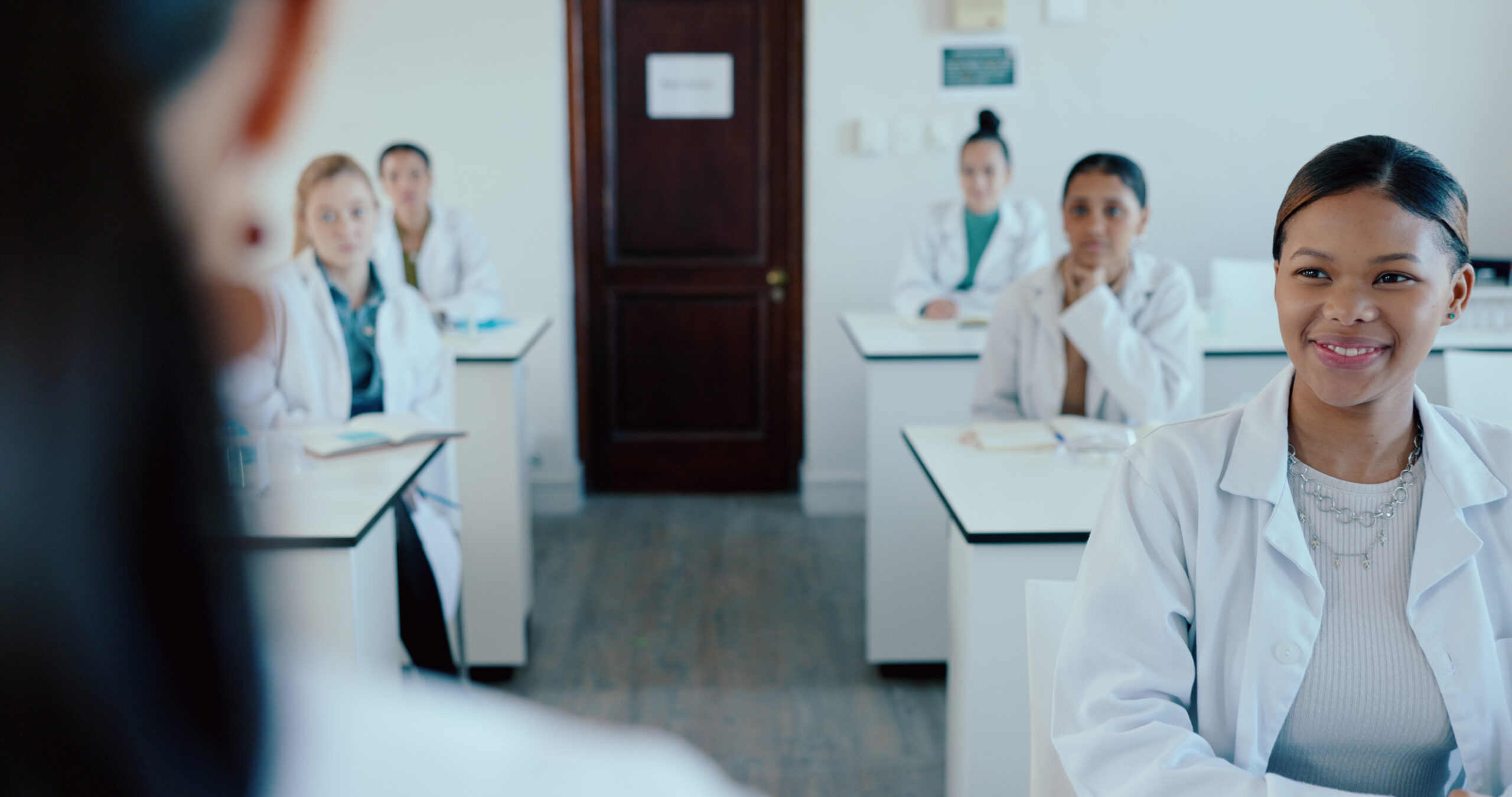 Female pharmacy students wearing white coats sit in a classroom.