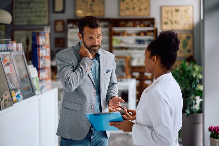 An adult male holds his neck in discomfort while seeking advice from a female pharmacist.