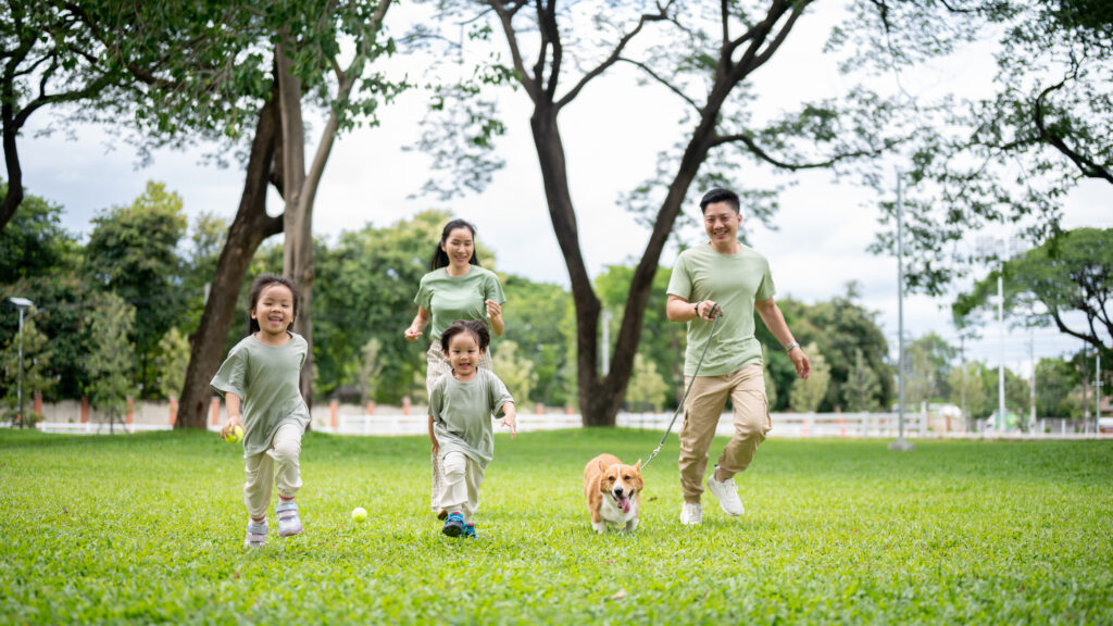 A young couple run in the park with two small children and a small dog. 