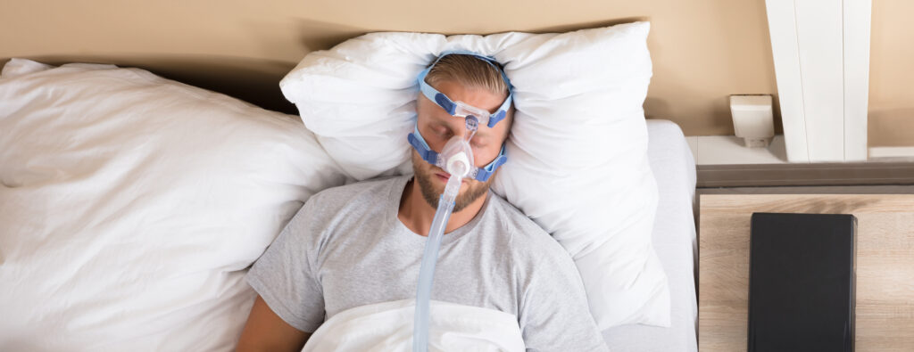 A man lays in bed sleeping wearing a CPAP machine. 