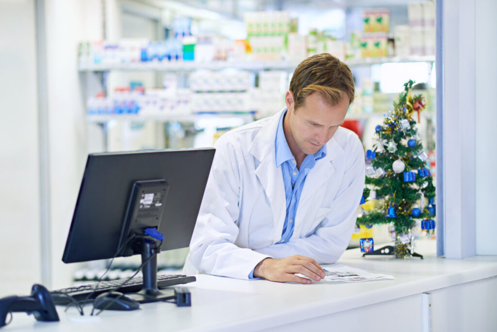 Un pharmacien se tient debout derrière le comptoir, en train de lire des documents, avec un petit sapin de Noël à côté de lui.