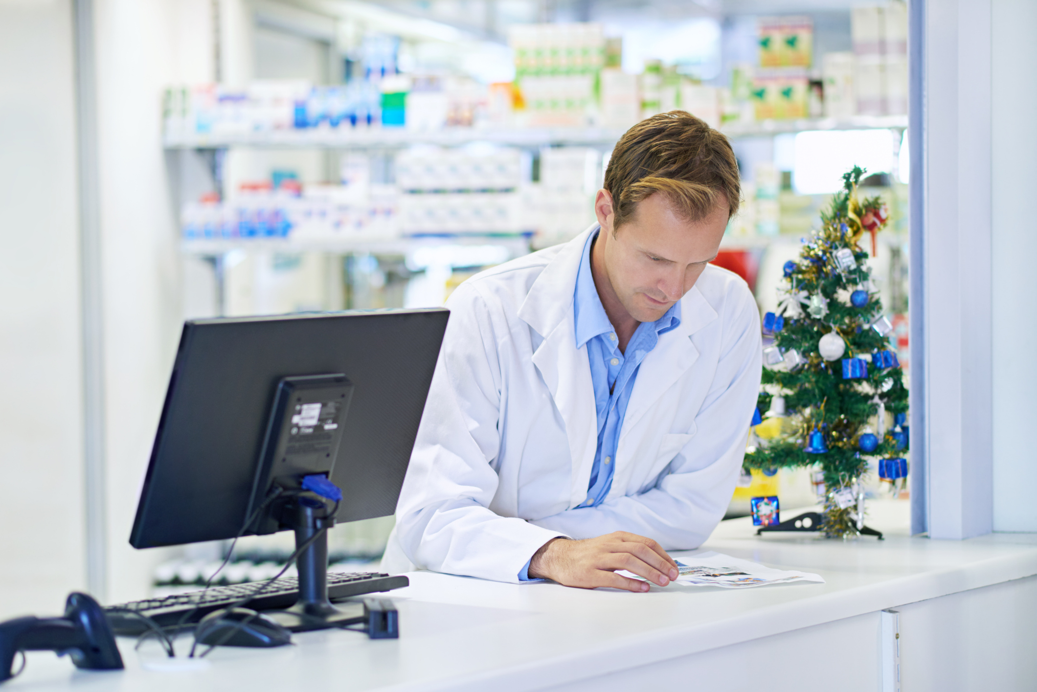 Un pharmacien se tient debout derrière le comptoir, en train de lire des documents, avec un petit sapin de Noël à côté de lui.