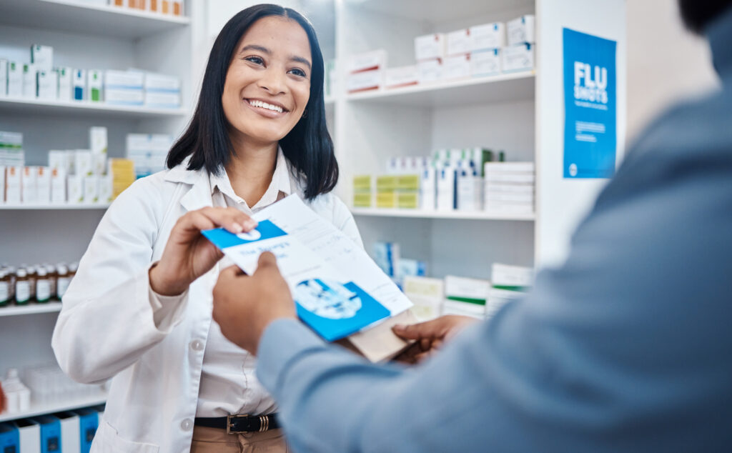 At the pharmacy counter, a female pharmacist smiles as she gives a male patient a brochure.