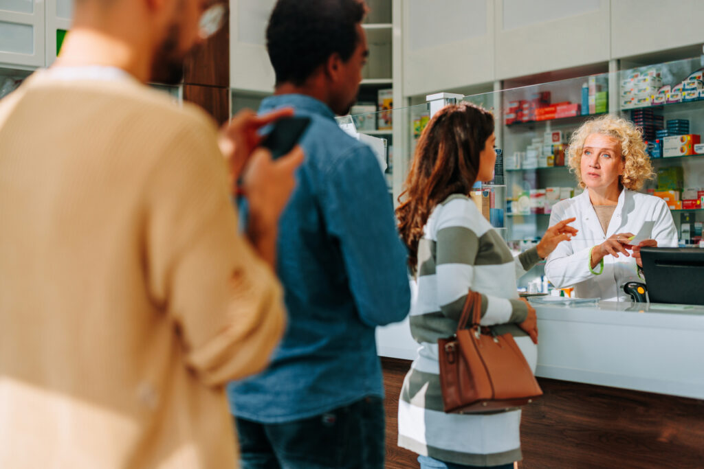 A female pharmacist helps a patient at the counter while two other patients wait in line.